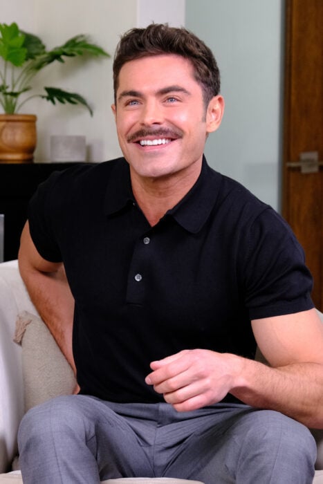 Photograph of Zac Efron sitting on a sofa with a mustache 