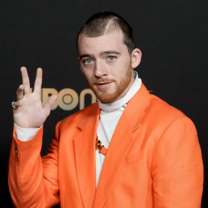 Angus Cloud vestido con un llamativo saco naranja y una camisa de cuello de tortuga blanca, saluda a las cámaras en una alfombra roja