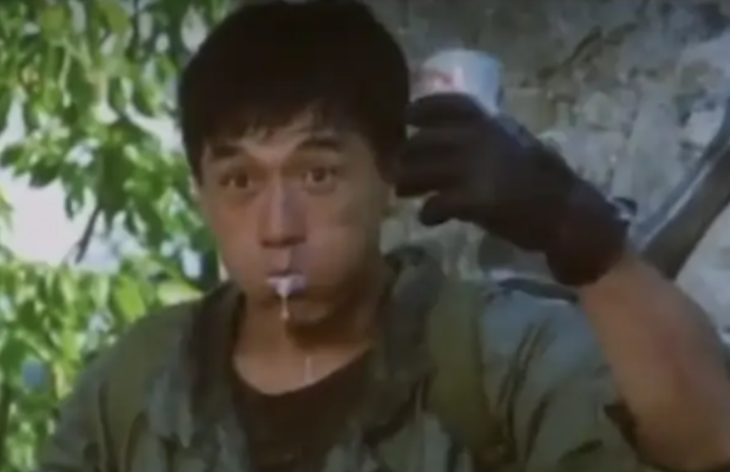 Jackie Chan bebe una cerveza que sostiene en una mano dejando escapar la espuma por la boca lleva guantes negros y un overol verde