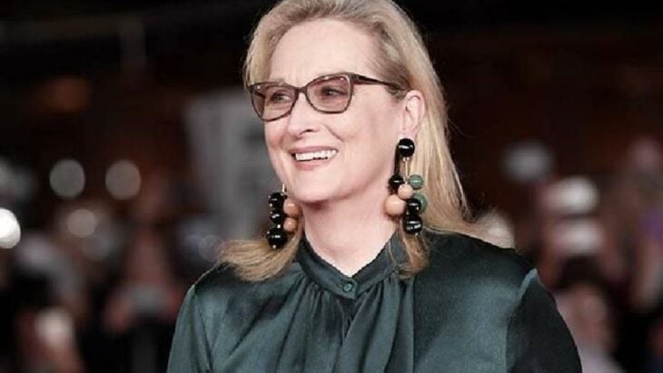 Meryl Streep con el cabello rubio suelto y largo luce unos aretes gigantes de color verde con negro lleva también una blusa verde oscuro