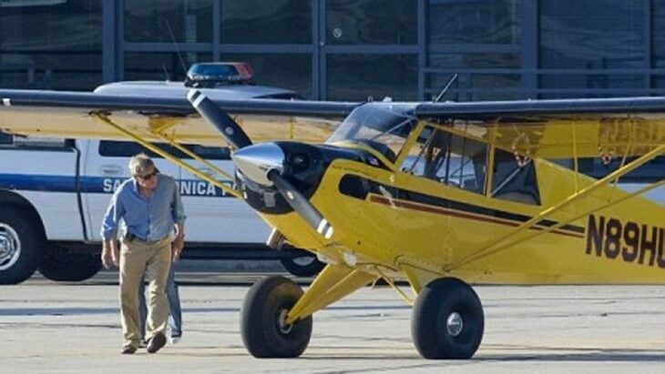 Harrison Ford revisando su avioneta amarilla para pilotearla lleva un pantalón café y camisa azul cielo