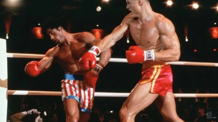 Sylvester Stallone en una pelea de box esta en el cuadrilátero recibiendo un puñetazo de su contrincante ambos visten shorts 