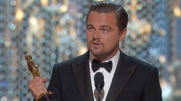 Leonardo DiCaprio agradeciendo un premio Oscar, esta dando un discurso en el micrófono viste un smoking negro con moño a juego
