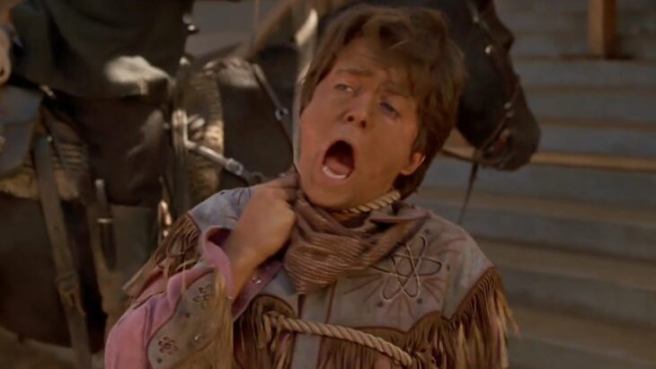 Michael J. Fox lleva una soga al cuello y esta haciendo un gesto de incomodidad pues simula que está siendo ahorcado viste con ropa típica del oeste