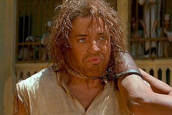 Brendan Fraser impartiendo realismo a una escena donde aparece con una soga al cuello simulando que es ahorcado lleva el cabello medio largo y despeinado con una camisa sucia color crema