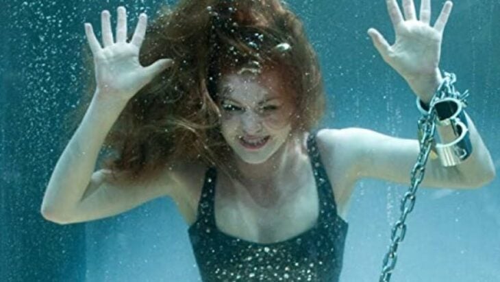 Isla Fisher dentro de un recipiente transparente lleno de agua y amarrada con cadenas lleva un traje de baño negro con pedrería