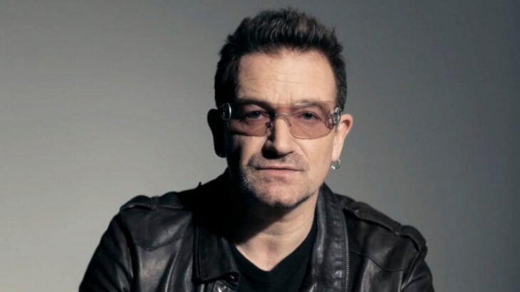 cantante Bono posando para la cámara con lentes semi transparentes una chamarra negra y playera negra trae un arete en una oreja