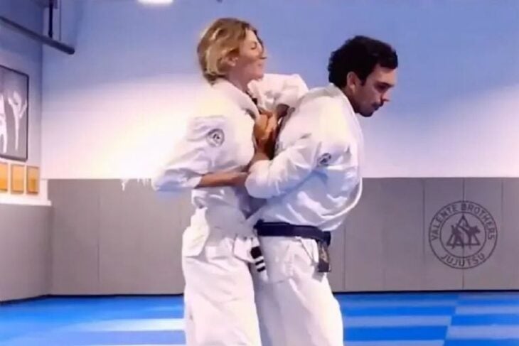Gisele Bundchen esá peleando en su clase de jiujitsu con su entrenador ella le practica una llave que mantiene inmovilizados los brazos del supuesto contrincante ambos llevan trajes blancos de karate