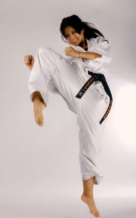 Courtney Cox está practicando una patada voladora lleva un traje blanco de karate y el cabello medio suelto una cinta negra le anuda la cintura