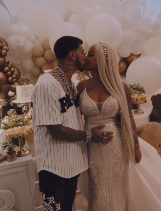Anuel AA and his wife Yailin The most viral girl at her daughter's gender reveal party, she wears a long white evening dress, her platinum blonde hair is loose and long, he wears a white baseball player type shirt with black stripes