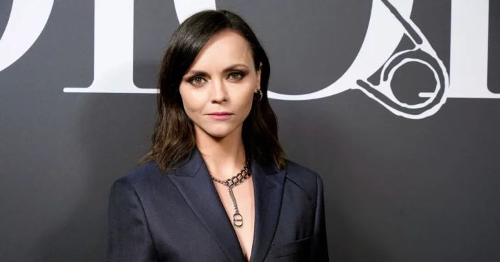 actress Christina Ricci posing for the cameras on a red carpet, she is wearing a navy blue tailored suit and a thick gold choker, her makeup is natural and her hair is in loose waves to shoulder length