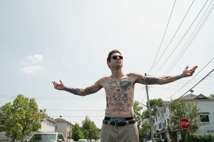Pete Davidson posando con los brazos abiertos sin camisa a la mitad de una calle con lentes de sol mostrando los tatuajes de su pecho 