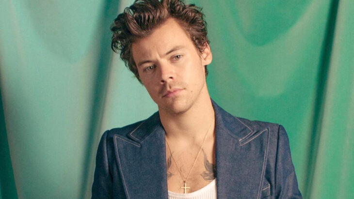 Harry Styles luciendo un saco azul de mezclilla con una playerita blanca lleva en el pecho un crucifijo de oro y el cabello ondulado y castaño