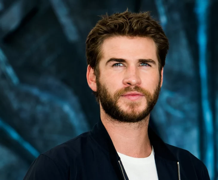 Liam Hemsworth en una sesión de fotos lleva un saco negro con una playera blanca resalta el azul de sus ojos por una pared de fondo oscura lleva barba y bigote