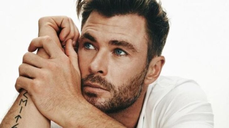 Chris Hemsworth posando en una sesión de fotos lleva una playera blanca y se lleva las manos a la cara dejando ver un tatuaje en su brazo derecho