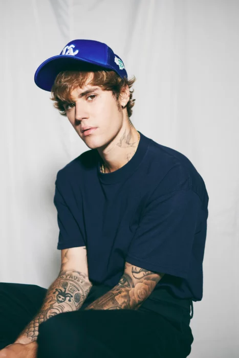 Justin Bieber sentado para una sesión de fotos viste todo en azul marino y lleva una gorra azul se aprecian los tatuajes en sus brazos