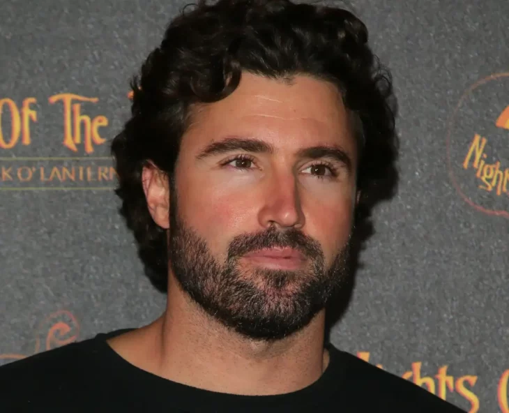 Brody Jenner esta parado frente a las cámaras en un evento lleva una sencilla camisa negra el cabello ondulado y una crecida barba y bigote