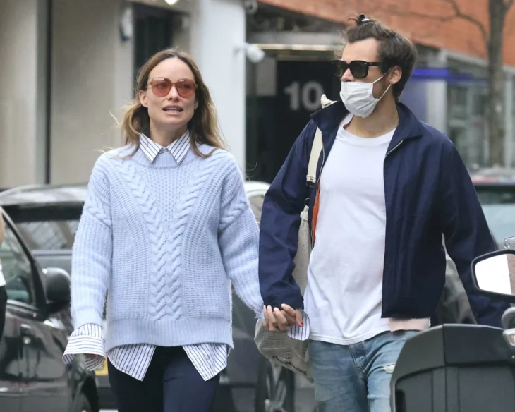 Harry Styles y Olivia Wilde caminando tomados de la mano, ella trae un sueter azul cielo con una camisa de rayas en blanco con azul él trae un jeans de mezclilla azul una camiseta blanca y una chamarra azul marino ambos usan gafas para sol