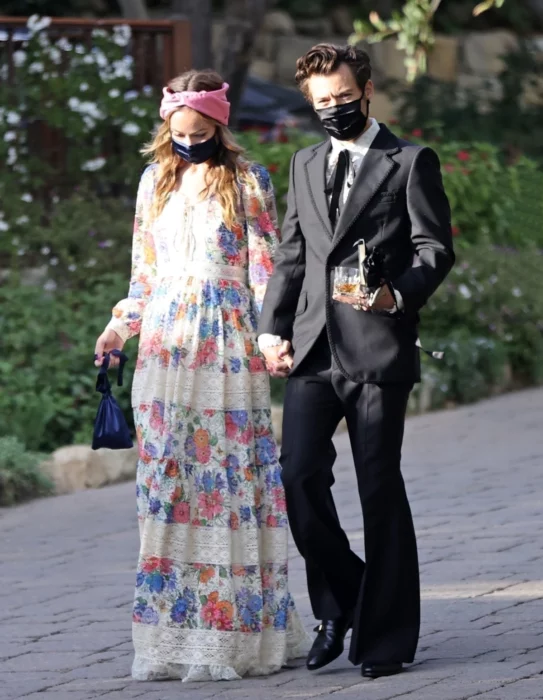 Harry Styles y Olivia Wilde caminando tomados de la mano, ella lleva un vestido casual largo con estampado floreado y una banda rosa sobre la cabeza, e´l porta un traje negro son camisa blnca y sorbata con forma de moño en negro, ambos usan cubrebocas negros