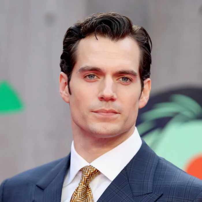 Henry Cavill dressed in elegant navy blue suit and gold tie, has short black hair and shows no beard or mustache
