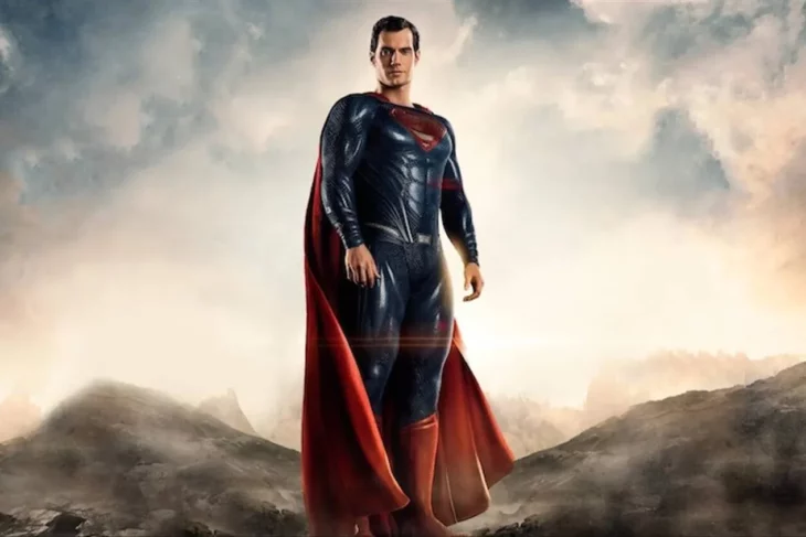 Superman character played by actor Henry Cavill wears a blue suit with red boots and cape on his chest he wears the letter S he is standing on some mountains with the sky behind