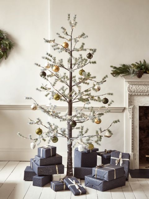árbol navideño con ramas secas con cajas de regalos en color gris debajo a lado de una chimenea en la sala de una casa 