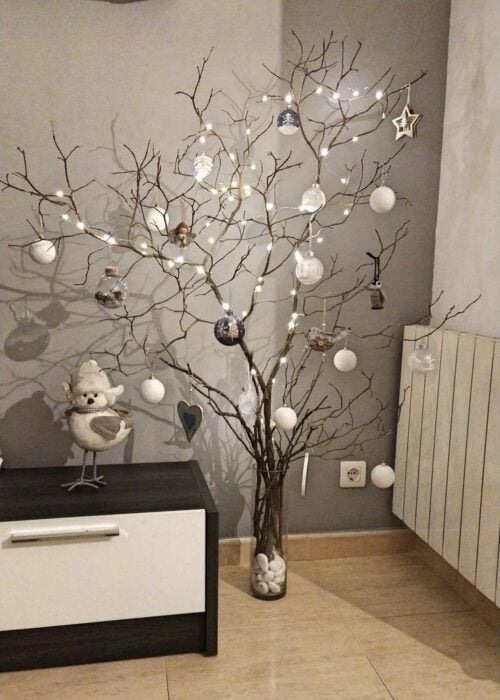 árbol de navidad hecho con ramas secas en la sala de una casa decorado con un mono de nieve y esferas de color blanco 