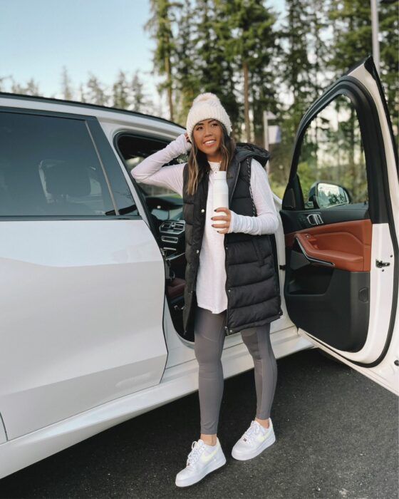 foto de una chica posando recargada en una camioneta rodeada de árboles usando un chaleco holgado y un gorro blanco 