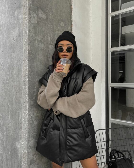 foto de una mujer recargada en una pared tomándose un café usando un chaleco holgado en color negro y un gorro negro 