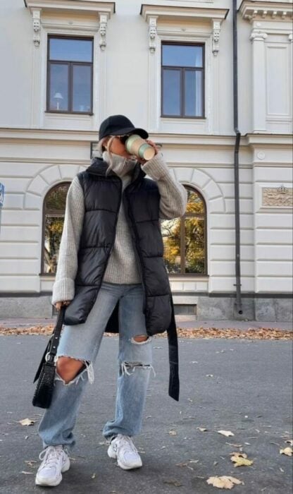chica con jeans rotos, un chaleco oversized tomando café a la mitad de una calle en una ciudad 