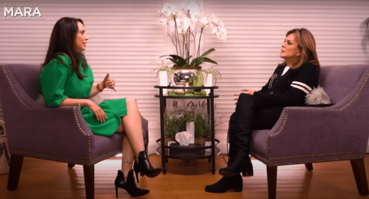 Dos mujeres hablando una frente a la otra, una con vestido verde y la otra vestida de negro, al fondo de la imagen hay unas flores blancas