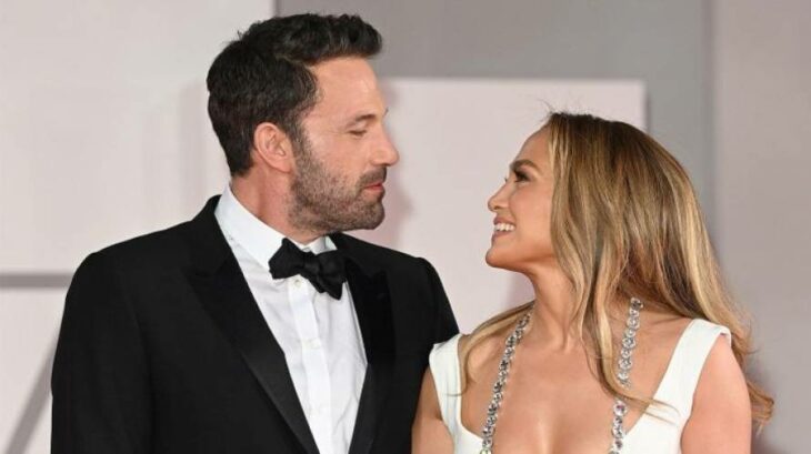 Jennifer López y Ben Affleck mirandose a los ojos ella lleva un vestido de noche blanco de tirantes y un collar de brillantes lleva el cabello suelto en tono rubio peinado con ondas él luce un smoking negro con camisa blanca y moño también negro 