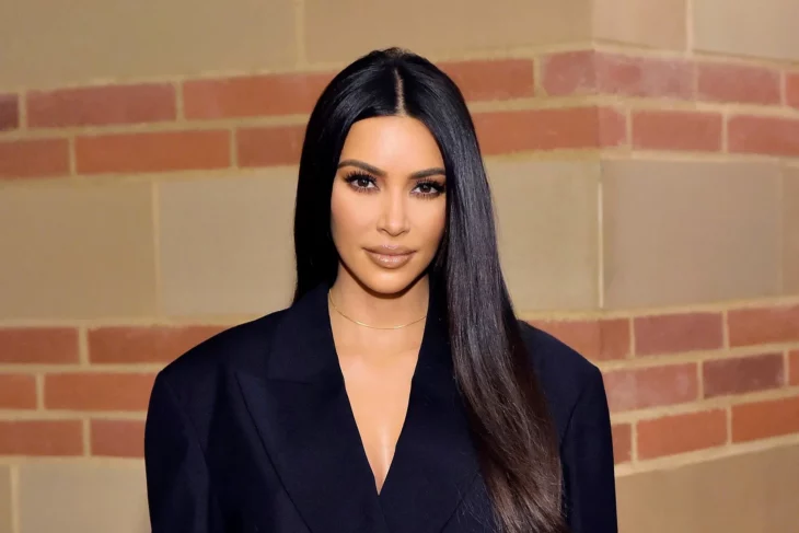 Kim Kardashian posando para la cámara lleva un traje sastre negro sin blusa abajo el cabello lo trae lacio y largo su maquillaje es discreto