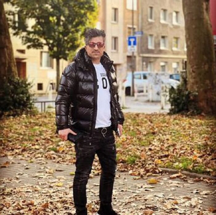 hombre posando en un parque público con cahammra y pantalones negros, lentes oscuros y playera blanca