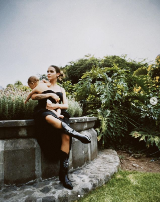 Yuya posando con su bebe en brazos sentada sobre una fuente de cantera lleva un vestido largo negro y con éste cubre a su pequeño hijo