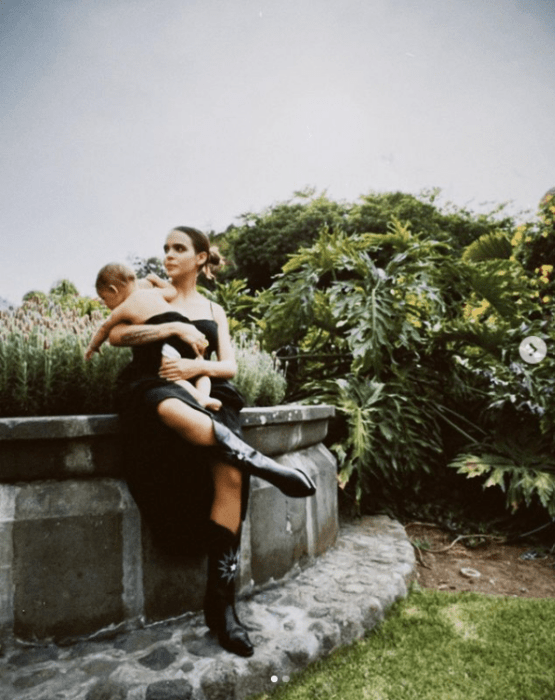 Yuya posing with her baby in her arms sitting on a quarry fountain wears a long black dress and covers her little son with it