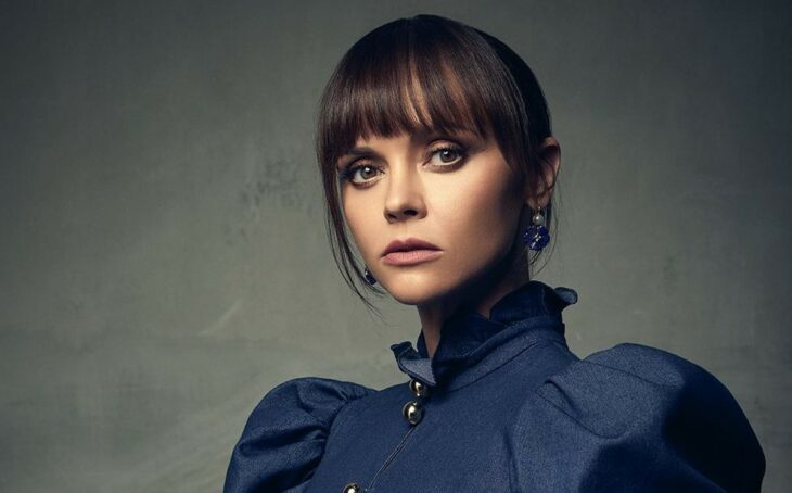 Christina Ricci wearing a navy blue blouse with thick sleeves and a high neckline, her hair is tied up in a bow and long fringe that covers her forehead