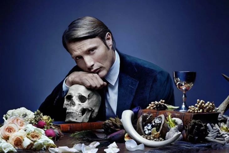 mads mikkelsen en su personaje de Hannibal, sentado en una mesa recargado en un cráneo