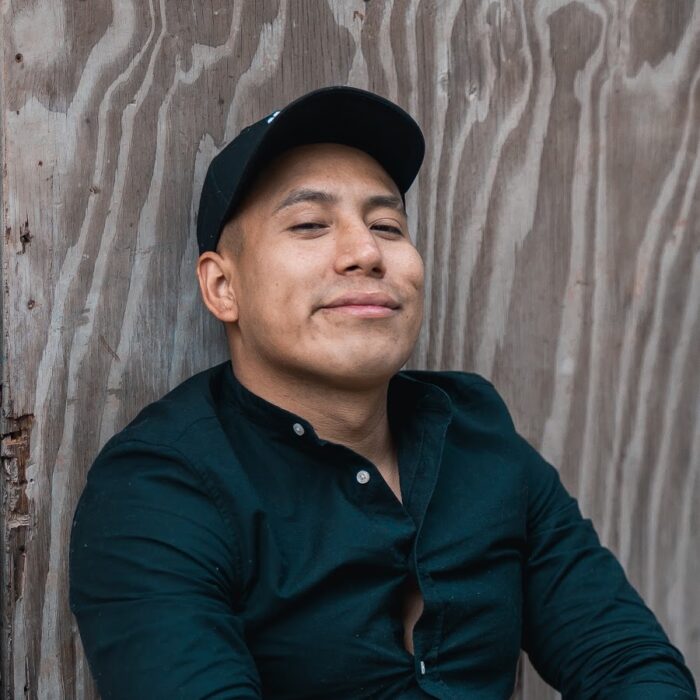 hombre moreno vestido con una camisa negra y gorra negra, recargado en una pared de madera gris posando para la foto 