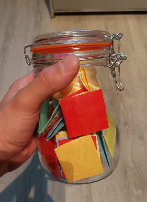 mano de un chico sosteniendo un frasco de vidrio con papeles de colores dentro 