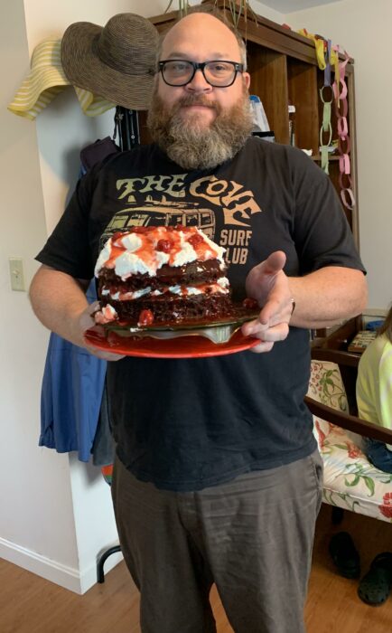 foto de un hombre vestido de negro sosteniendo en sus manos un plato con un pastel selva negra que hizo para su novia 