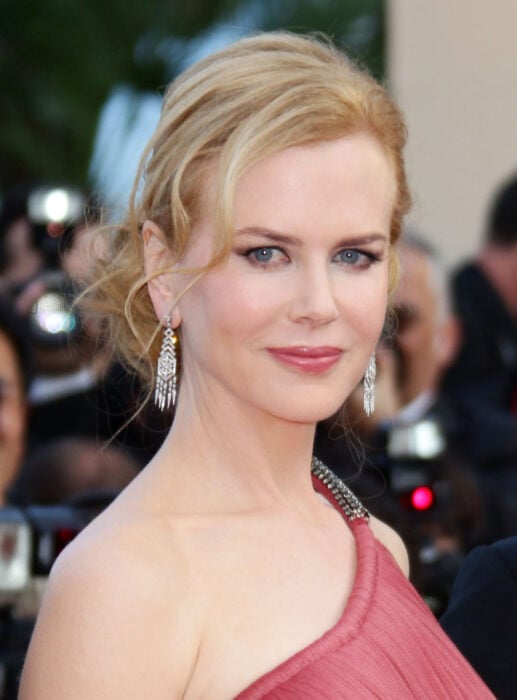 Nicole Kidman en una alfombra roja de una entrega de premios lleva un vestido de noche en tono rojo oscuro y unos pendientes de diamantes largos su peinado es un chongo recogido con mechones sueltos su maquillaje es marcado