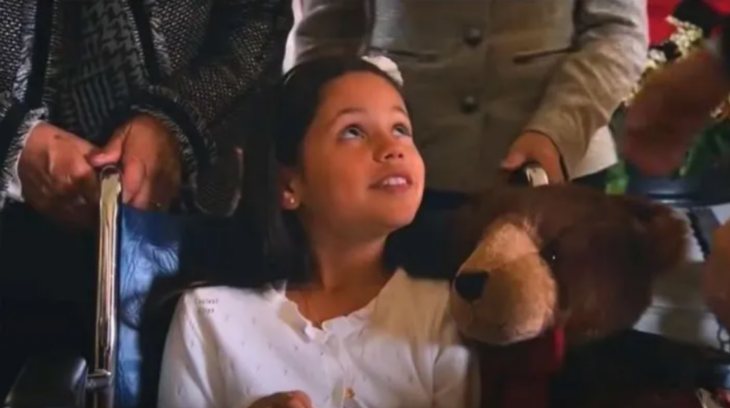 Jenna Ortega lleva un osito de peluche café esta interpretando un papel a sus 9 años lleva un blusa blanca y esta rodeada de personas