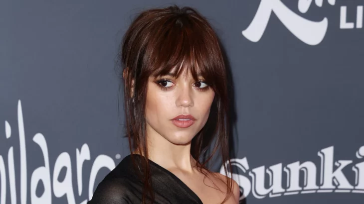 Jenna Ortega en una alfombra roja lleva un vestido negro de noche y un peinado de chongo con mechones sueltos el maquillaje es discreto 