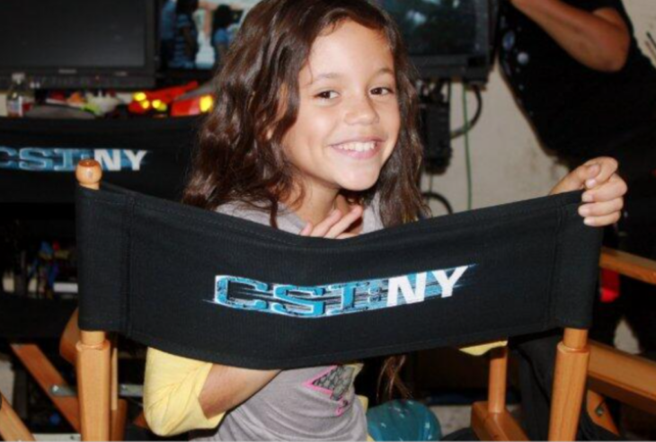 Jenna Ortega en una silla con el logotipo de CSI NY donde la chica participó tiene alrededor de nueve años lleva el cabello suelto y trae una sudadera gris con mangas amarillas