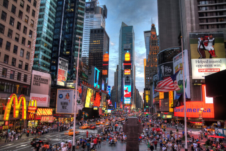 imagen de la ciudad de Nueva York esta anocheciendo y las luces de la ciudad están encendidas se ven los rascacielos y ríos de gente caminando 