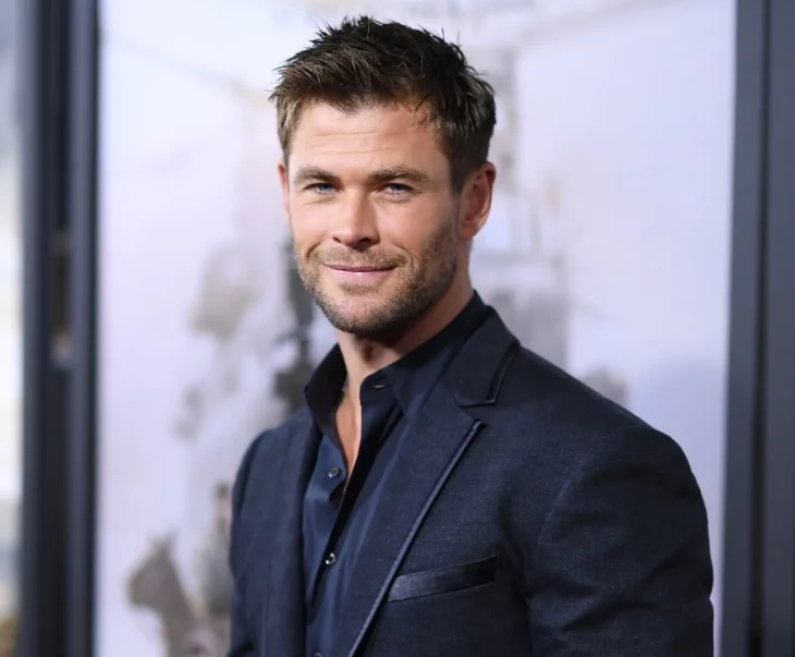 Chris Hemsworth posing for the cameras, he wears a jacket and black shirt without a tie, he has a smile on his face, his hair is short and a little disheveled 
