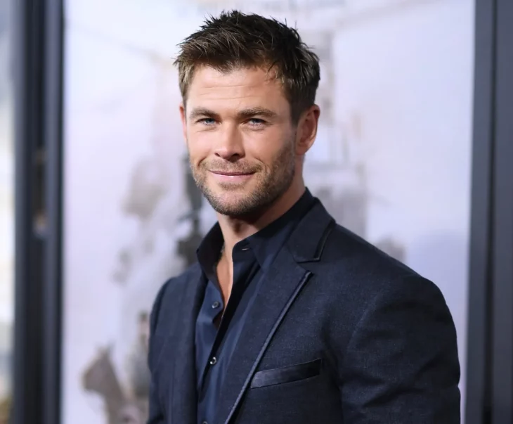 Chris Hemsworth posando para las cámaras lleva un saco y camisa negra sin corbata luce un gesto de sonrisa en su rostro lleva el cabello corto y un poco desaliñado 