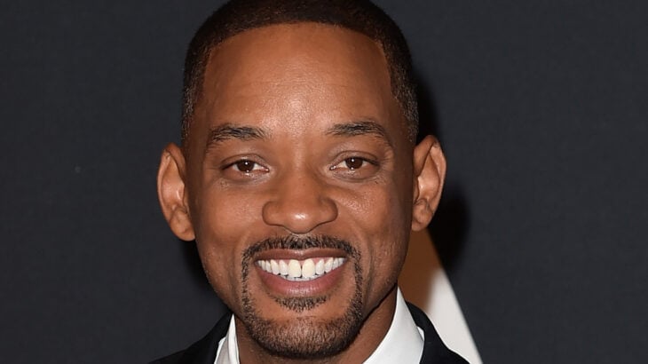 Will Smith smiles in an image where he can be seen standing on a red carpet wearing a black jacket and a white shirt 