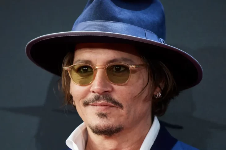 Johnny Depp luciendo un sombrero de gamuza azul marino con lentes redondos de sol lleva un arete en la oreja izquierda y una camisa de cuello blanco